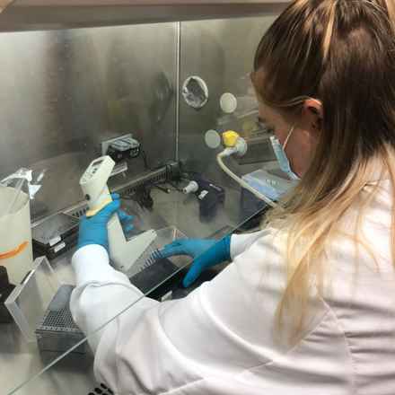 Elisha Griffin in the lab wearing a white lab coat, a face mask, and blue gloves, holding a scanner for an experiment